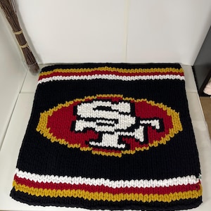 May include: A black, white, red, and gold knitted throw rug with the San Francisco 49ers logo in the center.