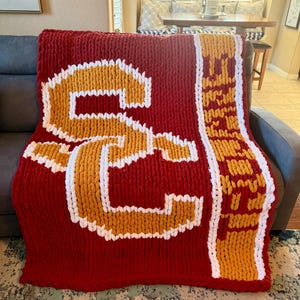 May include: A red, white, and gold knitted blanket with a large "S" in the center. The blanket is made of chunky yarn and has a soft, fuzzy texture.