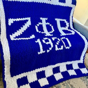 May include: A large, chunky knit blanket in royal blue with white accents. The blanket features the Greek letters ZΦB and the number 1920 in white. The border has a white and blue checkerboard pattern.