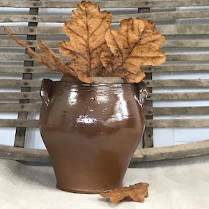 Könnte beinhalten: Ein brauner Keramiktopf mit zwei Henkeln und einer glasierten Oberfläche. Der Topf ist mit getrockneten Blättern gefüllt.