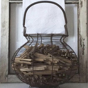 Könnte beinhalten: Ein rustikaler Drahtkorb, gefüllt mit Holzanzündern. Der Korb hat einen gebogenen Griff und steht auf kleinen Füßen. Ein weißes Tuch ist hinter dem Korb zu sehen, vor einem verwitterten, weiß gestrichenen Fensterrahmen.