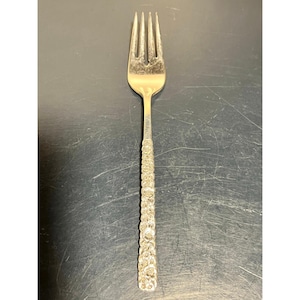May include: A silver fork with four tines and a textured handle. The handle features a repeating floral pattern. The fork is made of a shiny, metallic material and is designed for eating.