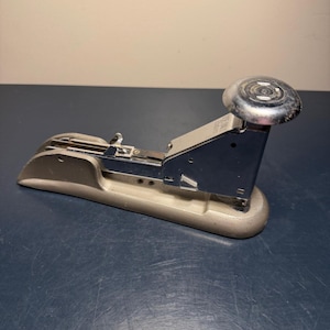 May include: A vintage silver and blue stapler with a rounded top and a curved base. The stapler is made of metal and has a classic design, suitable for office or home use. The stapler is shown on a dark blue surface.
