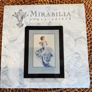 May include: Mirabilia cross-stitch kit featuring a framed image of a woman in a blue gown. The kit includes a black frame and is set against a background with white rose patterns. The Mirabilia logo is visible.