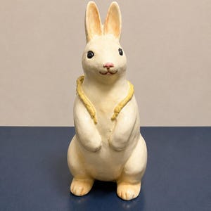 May include: A cream-colored ceramic rabbit figurine standing upright with its front paws tucked against its chest. The rabbit features long ears with light orange shading, black eyes, a pink nose, and a yellow textured collar around its neck.
