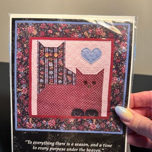 May include: A greeting card featuring a quilted design of two stylized cats. The cats are in shades of red and blue, with a blue heart. The card has a floral border and includes the text "To everything there is a season, and a time to every purpose under the heaven." Ecclesiastes 3:1.