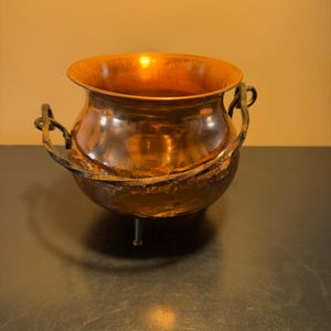 May include: A small, round, hammered copper cauldron with a wide, flared rim. The vessel features a dark, textured metal swing handle attached to two side loops and rests on three short, thin, dark metal legs for stability.