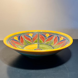 May include: A large, decorative ceramic bowl with a wide, shallow design. The bowl features a vibrant, hand-painted floral pattern in red, yellow, blue, and green against a yellow rim. The bowl's exterior is a dark teal color.