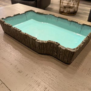 May include: Rectangular ceramic tray with a turquoise interior and a textured, brown exterior resembling tree bark. The tray sits on a light brown wooden surface. The tray's edges have a rough, uneven design.
