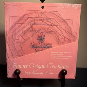 May include: A set of clear acrylic Flower Origami Templates by Kumiko Sudo in pink packaging. The packaging features the text Flower Origami Templates, Perfect Circles, Squares, Octagons, and Pentagons-Every Time!, and Three Sizes on Each All-in-One Template.