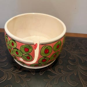 May include: A ceramic bowl with a pink exterior, decorated with green and red olive designs. The bowl has a white interior and a small notch on one side. A heart is painted on the side of the bowl.