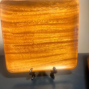 May include: A square wooden serving tray with rounded corners. The tray has a natural wood grain pattern in shades of brown and gold. It is displayed on a black metal stand, suitable for serving appetizers or displaying decorative items.