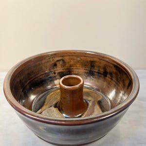 May include: A brown ceramic bundt pan with a central tube. The pan has a glossy finish with shades of brown, tan, and black. The rim and tube are a darker brown. The pan is designed for baking cakes.