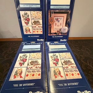 May include: Four Bucilla cross-stitch kits are displayed, each in a dark blue package. The kits feature the Zweigart brand and include various cross-stitch patterns. The packages display the text "FEEL THE DIFFERENCE" and include images of floral and animal designs.