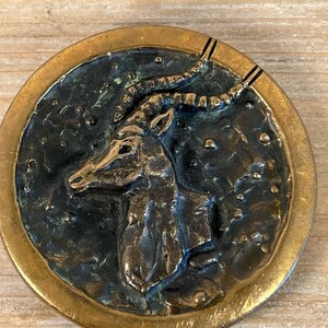 May include: A round, brass-colored button with a raised, detailed image of an antelope's head. The button has a textured, dark blue background with a gold rim.