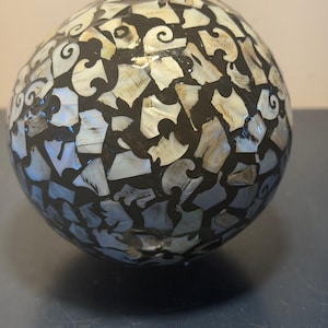 May include: A decorative sphere with a mosaic design. The surface is covered in irregular pieces of iridescent shell and dark, possibly black, material. The shell pieces have a pearlescent sheen, and the sphere is round.