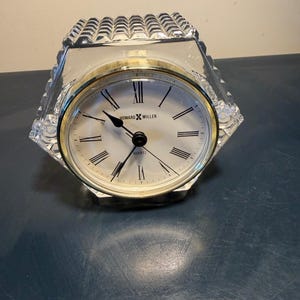 May include: A clear, faceted quartz clock with a gold-tone bezel and a white face. The clock features black Roman numerals and hands, with the brand name "HOWARD MILLER" visible on the face. The clock is set on a dark blue surface.