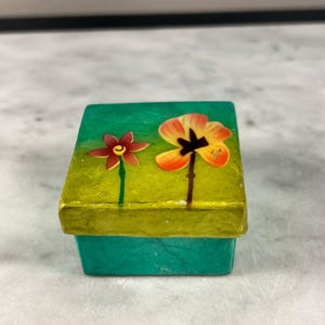 May include: A small, square box with a teal base and a yellow-green lid. The lid is decorated with two stylized flowers in shades of red, orange, and yellow. The box appears to be handmade and has a textured surface.