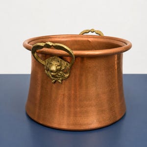 May include: A round copper pot with a smooth, metallic finish and a wide, rolled rim. The vessel features two decorative brass handles, each shaped like a lion head with a ring, attached to opposite sides of the exterior wall of the container.