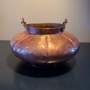 May include: A round, hammered copper bowl with a wide, bulbous body and a smooth, cylindrical neck. The vessel features two small, circular metal loops attached to the rim on opposite sides, designed for a handle or hanging mechanism.