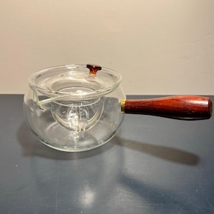 May include: A clear glass teapot with a wooden handle and lid. The teapot has a round body and a built-in infuser. The handle is made of dark wood and is attached to the pot with a brass fitting. The lid is also made of wood.