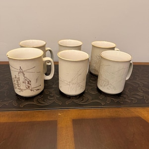 May include: Set of six beige ceramic mugs with landscape illustrations. Each mug features a different design, including a lighthouse, a bridge, and mountain scenery. The mugs have a simple, classic design with a light brown rim and handle.