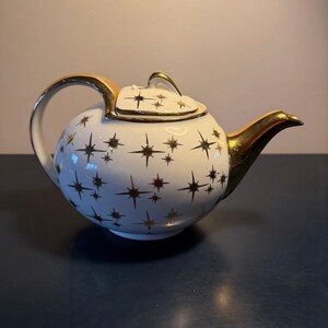 May include: White ceramic teapot with a gold handle, spout, and trim. The teapot is decorated with a pattern of gold starbursts. The lid also has gold trim and a small handle. A classic tea accessory.
