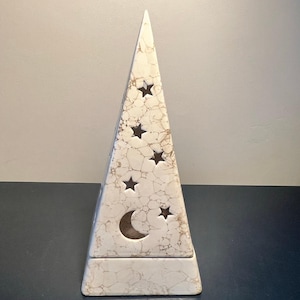 May include: A triangular ceramic luminary featuring a cream-colored base with a brown marbled crackle pattern. The surface is perforated with cutouts of a crescent moon and several five-pointed stars arranged vertically along the front face.