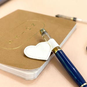 May include: A blue and gold fountain pen with a white heart-shaped charm rests on a brown notebook. Several black pens are arranged nearby. The notebook has gold embossed text. The background includes a laptop and a light-colored surface.