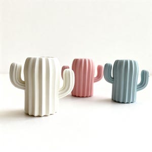 May include: Three ceramic cactus-shaped planters in white, pink, and blue. Each planter has a ribbed body and two curved arms, resembling a cactus. The planters are arranged on a white surface.