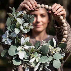 May include: A circular wreath featuring a ring of smooth, light-coloured wooden beads. The lower portion of the wreath is adorned with a dense arrangement of sage green eucalyptus leaves and soft, fuzzy, light green lamb's ear foliage.