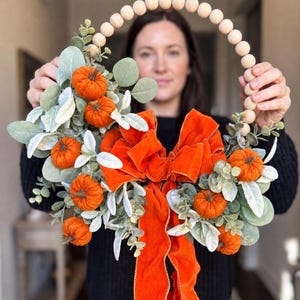May include: A circular wreath featuring a top arch of natural wooden beads, decorated with several small orange fabric pumpkins, green eucalyptus leaves, and a large, vibrant orange velvet bow with long, flowing tails hanging downward.