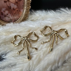 Puede incluir: Dos ganchos decorativos dorados con forma de lazo. Los ganchos son de metal y tienen un acabado brillante. Los ganchos están sobre una superficie blanca y esponjosa. Un marco de fotos ovalado dorado está en el fondo.