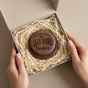 May include: A wooden baking tool set with the words "CAKE QUEEN" and "Jordan" engraved on it, presented in a gift box with shredded paper. The set includes a round, wooden piece with a ruler around the edge.