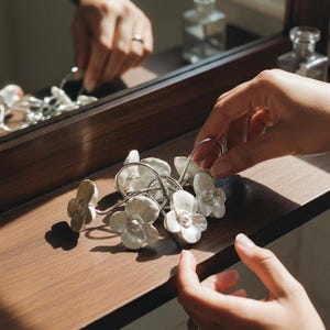 Pode incluir: Um conjunto de argolas de cortina brancas em cerâmica em forma de flor com laços de metal prateado. As argolas estão dispostas sobre uma superfície de madeira castanha escura, com um espelho e outros objetos decorativos no fundo. A imagem é bem iluminada.