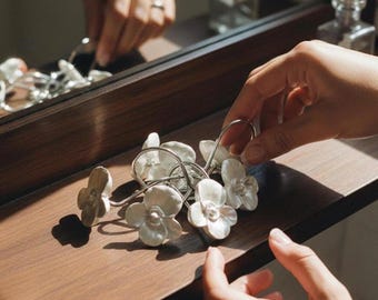 May include: A set of white ceramic flower-shaped curtain rings with silver metal loops. The rings are arranged on a dark brown wooden surface, with a mirror and other decorative items in the background. The image is well-lit, highlighting the details of the floral design.