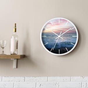 May include: White round clock with a picture of a seascape with a pink and orange sunset. The clock has white hands and black numbers.