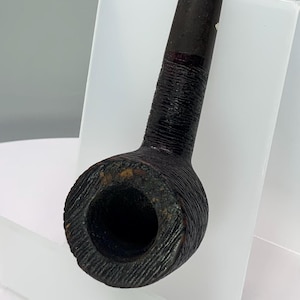 May include: A dark brown, vintage tobacco pipe with a textured bowl and stem. The pipe is made of wood and has a curved shape. The bowl is round with a large opening, and the stem is long and slender. The pipe is displayed on a white stand.