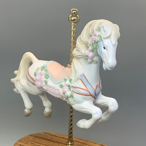 May include: A white porcelain carousel horse with pink roses and blue and orange ribbons. The horse is mounted on a wooden base with a gold pole and a gold "Fancy Land" logo.