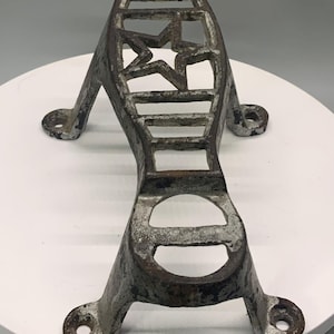 May include: A weathered, cast-iron footrest with a star-shaped cutout and rectangular openings. The metal has a distressed, aged appearance with visible rust and white paint. The footrest has a curved design with mounting holes for attachment.