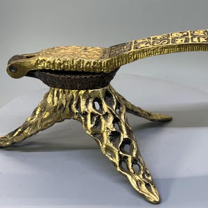 May include: A vintage brass nutcracker with a textured surface and a three-legged base. The handle features an embossed design. The nutcracker is gold-colored and appears to be made of metal.
