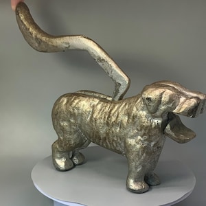 May include: A silver cast iron dog nutcracker with an open mouth and a lever on the back. The dog is standing on a white surface.