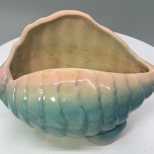 May include: A ceramic seashell planter with a gradient of colors. The exterior transitions from a light pink to a teal blue, while the interior is a light beige. The planter has a textured, ridged surface and a curved shape.