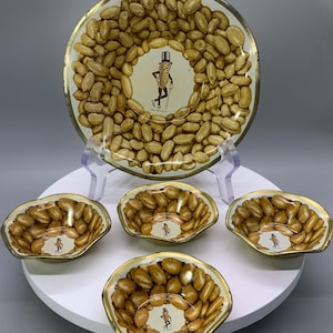 May include: Vintage Mr. Peanut serving set. The set includes a large round tray and four smaller serving dishes. The tray and dishes feature a peanut design with a gold trim. The center of each piece has a Mr. Peanut character.