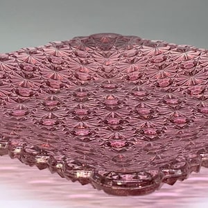 May include: A square purple glass plate featuring a raised geometric starburst pattern across its entire surface. The edges of the plate are scalloped and textured, creating a faceted appearance that catches light across the translucent material.