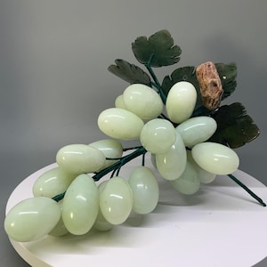 May include: A decorative cluster of pale green, smooth, oval-shaped stone grapes attached to a dark green wire stem. The arrangement features several dark green carved stone leaves and a single textured, brown, cylindrical stone piece.