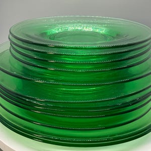 May include: A stack of emerald green glass plates. The plates are round with a slightly raised rim and a central indentation. The glass has a smooth, reflective surface, and the plates are stacked on top of each other.