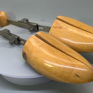May include: A pair of wooden shoe trees with metal arms. The shoe trees are light brown and have a smooth finish. The metal arms are silver and have a simple design.