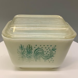 May include: Vintage Pyrex casserole dish with a clear glass lid. The off-white dish features a teal rooster and floral design. The square dish is approximately 7 inches wide and 3 inches tall.
