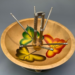May include: A wooden nut bowl with a painted leaf design in yellow, green, and red. The bowl holds a set of silver nutcrackers and picks. The bowl is light brown and has a smooth finish.